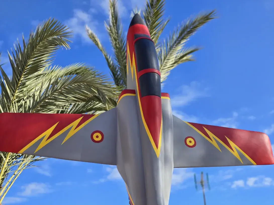 A sculpture of a jet plane painted in red points up at the sky, with palm trees behind